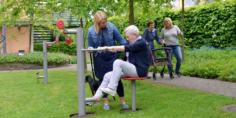 Eine Pflegerin hilft einer älteren Dame bei der Benutzung der Bewegungsgeräte im Garten des Seniorencentrums.